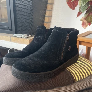 Svarta mockaboots från Rieker - Svarta boots från Rieker i mocka med extra bred passform. Skorna har dragkedja på sidan och en grov, platt sula för bra grepp. Perfekta för dig som vill ha en stilren och bekväm sko i äkta skinn till höst och vinter.