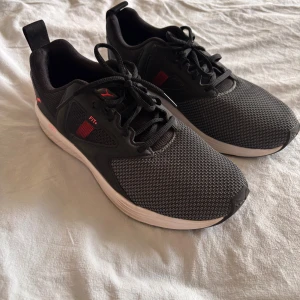 Svarta sneakers från Puma - Snygga svarta Puma sneakers med mesh-ovandel och vita sulor. Skorna har svarta snören, cerise Puma-loggor och detaljer samt en sportig design. Perfekta för dig som gillar stilrena och bekväma skor till vardags eller träning.