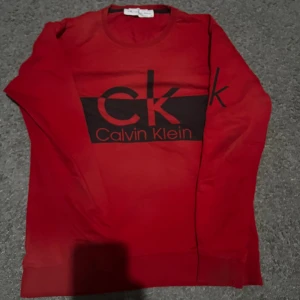 Röd Calvin Klein tröja med logga - Röd långärmad tröja från Calvin Klein Jeans med stor svart CK-logga och text på bröstet. Tröjan har rund halsringning och ribbade muddar. Tillverkad i mjuk bomull, perfekt för en avslappnad och stilren look.
