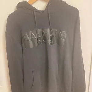  hoodie från Balmain Paris - Svart hoodie från Balmain Paris med stor logga i svart på bröstet. Trycket på bröstet har släppt vid kanter, annars felfri