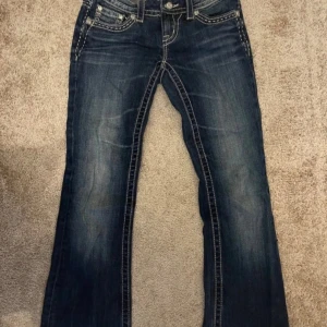 Miss Me bootcut jeans med detaljer - Snygga mörkblå bootcut jeans från Miss Me med kontrastsömmar, broderade bakfickor och glittriga knappar. Jeansen har låg midja och coola detaljer som metallmärke och dekorativa sömmar. Perfekta för dig som gillar statement-jeans med unik stil.