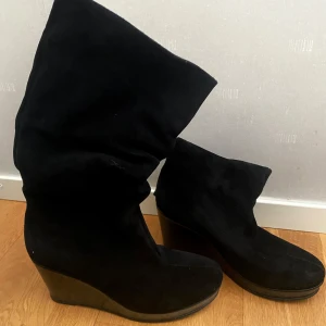 Svarta wedge boots i mocka - Säljer ett par svarta boots med kilklack i mocka. Skorna har en hög, slouchy skaft och plattformsula som ger extra höjd. Bekväma att gå i även under kalla vintrar och regnigt väder under hösten, 6cm i kilklacken. Använt sparsamt och tagit hand om väl! Storleken är 38 men passar även 37!