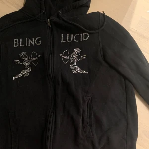 Svart hoodie från Bling Lucid  - Svart hoodie från Bling Lucid med dragkedja och huva. Framsidan har  strassdetaljer med texten 'BLING LUCID' och två änglar i strass. Endast testad. Pris går att diskutera🌸 sorlek M/L