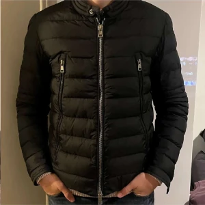 Svart Moncler pufferjacka herr - Snygg svart pufferjacka från Moncler med quiltad design och två bröstfickor med dragkedja. Jackan har ståkrage, lång ärm och Moncler-logga på vänster arm. Perfekt för dig som vill ha en clean och stilren look under höst och vinter.