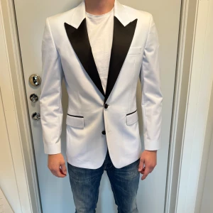 Vit kavaj med svarta slag - Säljer en stilren vit kavaj med breda svarta slag och svarta detaljer. Storleken är 46. Kavajen är använd 2 gånger 