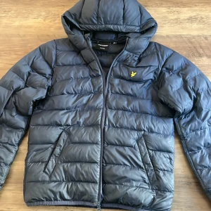 Mörkblå pufferjacka från Lyle & Scott - Snygg mörkblå pufferjacka från Lyle & Scott med huva och dragkedja. Jackan har det klassiska gula logobroderiet på bröstet och två snedställda fickor framtill. Perfekt för kyliga dagar och riktigt skön att bära.