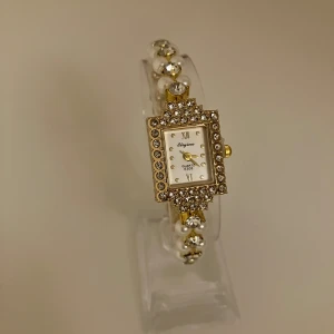 Guldig klocka med pärlor och stenar - Snygg klocka från Eliyina med fyrkantig urtavla, guldig ram och glittrande stenar runt om. Armbandet är dekorerat med vita pärlor och guldfärgade detaljer. Perfekt accessoar för dig som gillar bling och elegans.