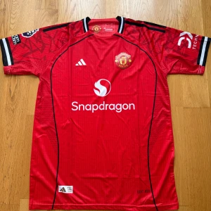 Manchester United röd matchtröja Adidas - Snygg Manchester United fotbollströja från Adidas i rött med svarta och vita detaljer. Tröjan har klubbmärke på bröstet, Snapdragon sponsortryck och korta ärmar. Tillverkad i lätt funktionsmaterial, perfekt för match eller träning. Storlek M