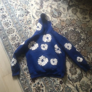 Blå hoodie från Denim Tears med blommor - Säljer en blå hoodie från Denim Tears med vita blommiga tryck över hela tröjan. Tröjan har huva, magficka och är gjord i 100% bomull. Perfekt för dig som vill sticka ut med en unik och snygg streetwear-look.