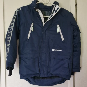 Blå vinterjacka från Wallride, stl 128 - Mörkblå vinterjacka från Wallride med vita detaljer och tryck på ena ärmen. Jackan har flera fickor med dragkedja, hög krage, reflekterande detalj bak och justerbara ärmslut. Perfekt för kalla dagar.. Man kan se slitage på jackans nederkant och vid ärmslutet (se bilder).