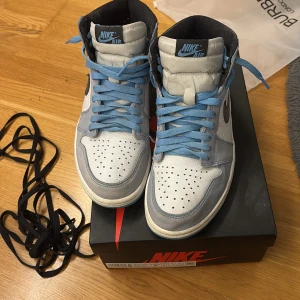Nike Air Jordan 1 High University Blue - Nike Air Jordan 1 High i University Blue med vit och ljusblå mocka, svarta detaljer och blå snören. Klassisk siluett med perforerad tå, svart swoosh och högt skaft. Perfekta för dig som vill sticka ut med en ikonisk sneaker. Får med svarta snören. Kan inte garantera äkthet