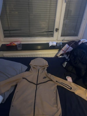 Nike tech - Snygg beige hoodie från Nike med hel dragkedja och svarta detaljer. Hoodien har huva och två sidofickor, samt Nike-logga på bröstet. Perfekt för en avslappnad och sportig stil. Säljer för behöver pengar helt nytt