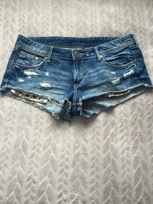 Blå low waist jeans shorts - Säljer ett par blå jeansshorts med slitna detaljer och fransig kant. Shortsen är i stl 40 men själv skulle jag nog säga att de sitter som 38. Shortsen har klassisk femficksmodell, låg midja och dragkedja framtill. Perfekta för sommaren! Pris går självklart att diskuteras 
