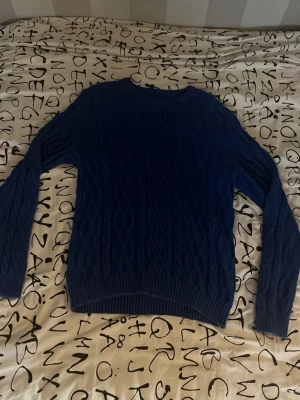 Blå kabelstickad tröja från Crocker - Säljer en blå kabelstickad tröja från Crocker. Tröjan har rund halsringning, långa ärmar och ribbade muddar vid ärmslut och nederkant. Perfekt för dig som gillar klassisk stil med en twist. Materialet känns mjukt och bekvämt.