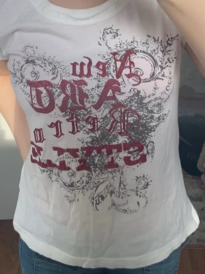 Vit t-shirt med tryck - Vit t-shirt med korta ärmar och tryck i röd glittertext och grått mönster framtill. Snygg och trendig design med dekorativa detaljer. 