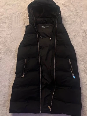 Dunväst  - Lång svart dunväst från Zara med huva och dragkedja framtill. Västen har två fickor med dragkedjor och justerbar dragsko vid huvan. Perfekt för hösten, vår och vinter
