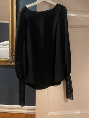 Svart blus med spetsdetaljer - Stilren svart blus med långa puffärmar och breda spetsmanschetter. Blusen har en rak passform och båtringad hals. Materialet är mjukt tyg med spetsdetaljer vid ärmsluten som ger en elegant touch.