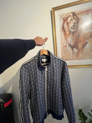 Blåvit mönstrad  Lacoste - Säljer en blåvit träningsväst från Lacoste med hel dragkedja och hög krage. Västen har ett geometriskt mönster, vita ränder längs ärmarna och Lacoste-logga på bröstet. Perfekt för dig som gillar sportig och stilren look.