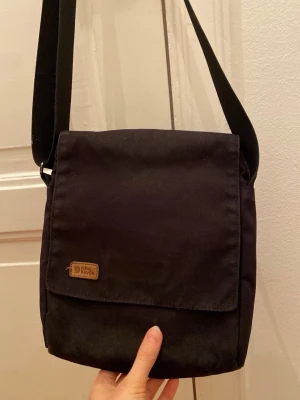 Svart axelväska från Fjällräven - En stilren svart axelväska från Fjällräven med justerbar axelrem och klassisk logga på framsidan. Väskan har en fyrkantig form och är tillverkad i slitstarkt tyg, perfekt för att bära dina viktigaste grejer.