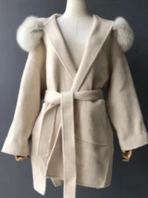 Beige pälsjacka med huva och bälte - Stilren beige pälsjacka med stor huva och fluffiga pälskanter. Jackan har knytskärp i midjan, stora fickor och en avslappnad passform. Perfekt för kalla dagar när du vill känna dig både varm och snygg. Äkta päls 