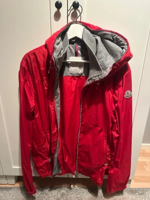 Moncler urville vindjacka med huva - Snygg röd vindjacka från Moncler med grått innerfoder och huva. Jackan har dragkedja framtill, två sidofickor och Moncler-logga på ärmen. Jackan är använd ett fåtal gånger. Skriv vid funderingar!