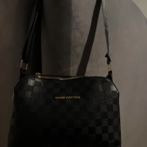 Svart fashion handväska - Snygg svart handväska med klassiskt rutigt mönster och guldfärgade detaljer. Väskan har justerbar axelrem och dragkedja med. Perfekt för dig som vill ha en stilren och lyxig accessoar.