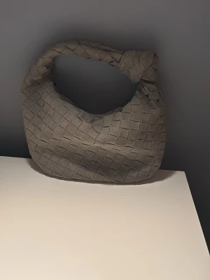 Grå flätad handväska i mocka - Trendig beige handväska med flätad design och rundad form. Väskan har ett brett handtag med knutdetalj och är tillverkad i mjuk mocka. Perfekt accessoar för dig som vill ha en stilren och modern look.