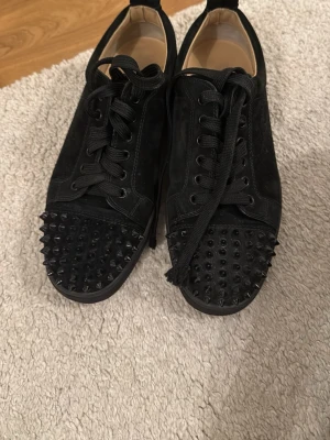 Svarta Louboutin  - Säljer ett par svarta sneakers från Christian Louboutin med klassisk röd sula och svarta nitar på tån. Skorna har snörning och är tillverkade i mocka för en lyxig känsla. Perfekta för dig som vill sticka ut med en edgy detalj.