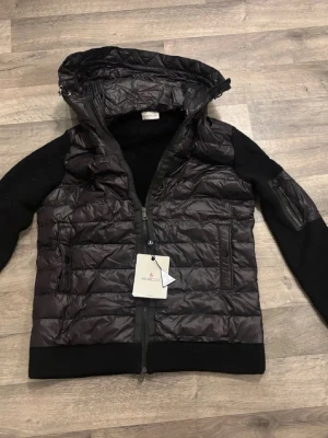 Svart dunjacka från Moncler med huva - Snygg svart dunjacka från Moncler med quiltad front och huva. Jackan har svarta stickade ärmar och detaljerad dragkedja framtill. Klassisk Moncler-logga på ärmen och praktiska fickor. Perfekt för kyliga dagar och riktigt stilren look.