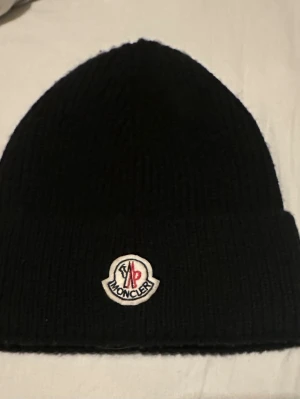Svart Moncler mössa i ull - Snygg svart ribbstickad mössa från Moncler med klassisk uppvikt kant och broderad logotyp framtill. Tillverkad i mjuk ull som håller dig varm och stilren under kalla dagar. Perfekt accessoar för dig som gillar exklusiva märken och enkel design.