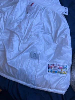 Vit dunjacka från Moncler - Säljer en snygg vit dunjacka från Moncler med quiltad design och dragkedja framtill. Jackan har två praktiska fickor med dragkedja och hög krage. Perfekt för kalla dagar och ger en clean look.