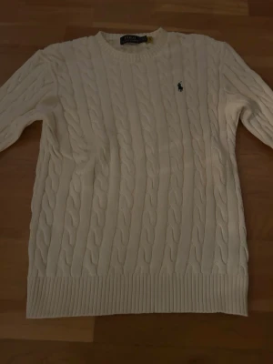 Vit kabelstickad tröja Polo Ralph Lauren - Kabelstickad vit tröja från Polo Ralph Lauren med rund halsringning och broderad logga på bröstet. Tröjan har ribbade muddar vid ärmslut och nederkant, perfekt för lager på lager. Klassisk och tidlös design.