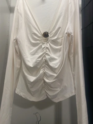 Vit långärmad topp med ringdetalj - Vit långärmad topp från DIVIDED med djup v-ringning och snygg ringdetalj framtill. Toppen har rynkade detaljer längs mitten som ger en cool och trendig look. Perfekt att styla med jeans eller kjol. 