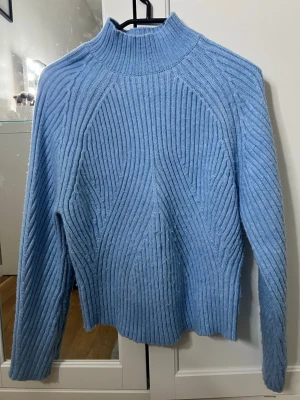 Ljusblå stickad tröja med struktur - Ljusblå stickad tröja med hög krage och snyggt strukturerat mönster. Tröjan har långa ärmar och ribbstickade detaljer som ger en modern look. Perfekt för att hålla värmen under kyliga dagar.