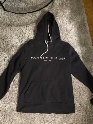 Svart hoodie från Tommy Hilfiger - Snygg svart hoodie från Tommy Hilfiger med klassisk logga och text 'EST. 1985' på bröstet. Hoodien har en stor ficka framtill och vita dragsnören i huvan. Perfekt för en avslappnad och trendig look.