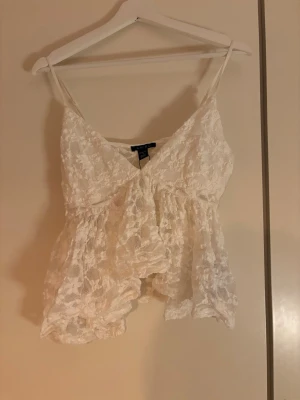 Vit spetslinne  - Söt vit linnetopp med tunna axelband och v-ringning. Toppen är täckt av blommig spets och har en luftig, flowy passform. Perfekt att styla med jeans eller kjol för en fräsch look.
