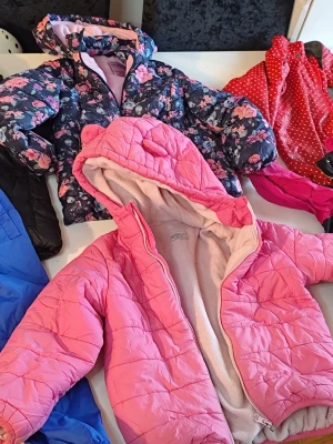 Barnklädespaket med jackor och regnbyxor stl 110/116 - Detta är ett paket med barnkläder. Det inkluderar jackor i olika färger och mönster, samt ett par blå regnbyxor. Jackorna har detaljer som huva och dragkedja. En av jackorna är rosa med öron på huvan, en annan är mörkblå med blommor(utan blixtlås), och en tredje är röd med vita prickar. Det finns även en svart jacka och en rosa jacka med svarta och limegröna detaljer(regnjacka).Du är varmt välkommen att kika på mina annonser.