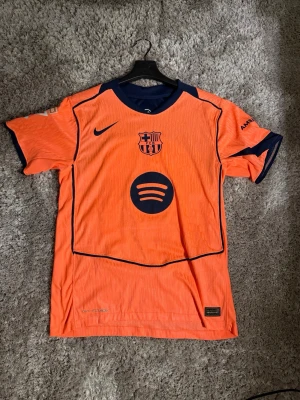 FC Barcelona matchtröja  - Snygg orange FC Barcelona matchtröja med mörkblå detaljer, Nike-logga och Spotify som sponsor på bröstet. Tröjan har F. De Jong och nummer 21 på ryggen samt UNHCR-logga. Tillverkad i lätt och ventilerande material, perfekt för fotboll eller träning.
