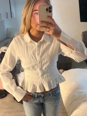 Vit blus med volang och krage - Snygg vit blus med klassisk krage, knäppning framtill och volang nertill. Blusen har långärmade ärmar och smockad midja som ger en feminin siluett. Perfekt att styla med jeans för en trendig look.