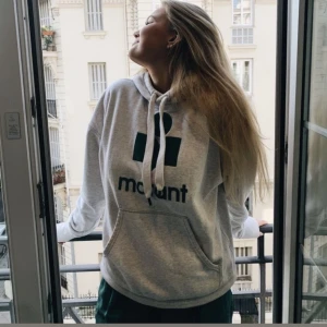 Grå hoodie från Isabel Marant - Snygg grå hoodie från Isabel Marant med svart logga framtill. Hoodien har huva med dragsko och en stor magficka. Tillverkad i mjukt bomullsmaterial och har en avslappnad passform. Perfekt för en chill och trendig look. Endast anbänd en gång!