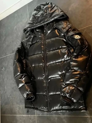 Svart M Fashion dunjacka med huva - Snygg svart dunjacka från M Fashion med glansig finish och huva. Jackan har dragkedja framtill, M fashion-logga på ärmen och quiltad design. Perfekt för kalla dagar och ger en riktigt clean look.