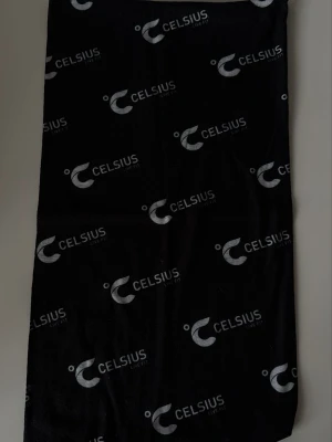 Svart halsvärmare med Celsius-logga - Snygg svart halsvärmare med upprepade vita Celsius-loggor tryckta över hela tyget. Perfekt accessoar för dig som gillar sportiga och trendiga detaljer. Tunn och smidig, lätt att styla till olika outfits.