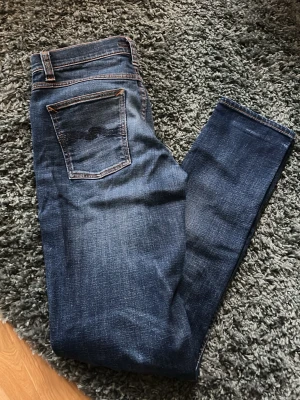 Nudie Jeans blå - Snygga mörkblå jeans med klassiska orange sömmar och en bakficka med vågig detalj. Jeansen har en rak passform och är tillverkade i slitstarkt denimtyg. Perfekta för dig som gillar en enkel men stilren look.