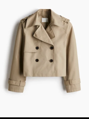 Beige croppad trenchcoat - Stilren beige croppad trenchcoat med dubbelknäppning och klassisk krage. Jackan har långa ärmar med spänndetaljer vid handlederna och axelklaffar. Perfekt för dig som vill ha en modern twist på en klassiker.