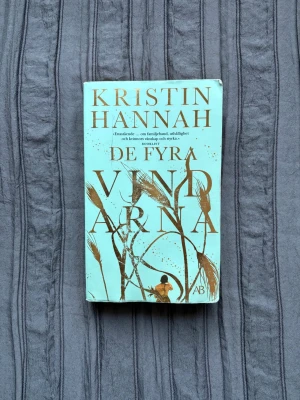 De fyra vindarna av Kristin Hannah - En roman av Kristin Hannah med turkos omslag och guldiga detaljer. Boken handlar om familjeband, uthållighet och kvinnors styrka under 1930-talets Amerika. Omslaget har illustrationer av vete och en person i siluett. Perfekt för dig som gillar gripande berättelser.