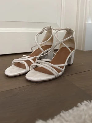 Vita pumps med remmar och blockklack - Säljer ett par vita pumps med tunna remmar och blockklack. Skorna har öppen tå och justerbart spänne vid ankeln. Snygg minimalistisk design som passar till många olika looks. 