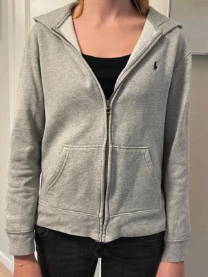 Ralph Lauren Hoodie L, 14-16 - Grå hoodie från Polo Ralph Lauren med  hästen i mörkblått, stl L/14-16, i fint skick. Storleken motsvarar XS.  Köpt på Kids Brand Store.  Mått Axel till axel 41 cm Längd från nacken ner 56cm Arm 55cm Bredd nedtill 47cm