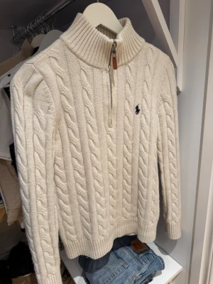 Vit kabelstickad tröja Polo Ralph Lauren - Kabelstickad vit tröja från Polo Ralph Lauren med halv dragkedja och hög krage. Tröjan har ribbade muddar och det klassiska mörkblå logobroderiet på bröstet. Tillverkad i mjukt material som ger en lyxig känsla och passar perfekt till jeans.