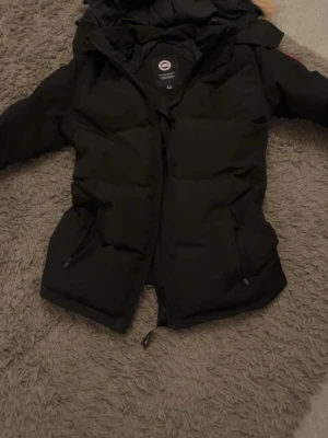 Svart dunjacka från Canada Goose - Säljer en svart dunjacka från Canada Goose med klassisk patch på ärmen. Jackan har dragkedja framtill, två fickor med dragkedja och en mysig huva med pälskant. Perfekt för kalla vinterdagar och riktigt snygg streetstil. Storlek m men passar även s 