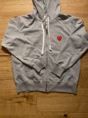 Grå hoodie från Comme des Garçons - Snygg grå zip-hoodie från Comme des Garçons Play med det ikoniska röda hjärtat med ögon broderat på bröstet. Hoodien har vita dragsnören, fickor fram och är tillverkad i mjuk bomullsblandning. Perfekt för en chill och trendig look.